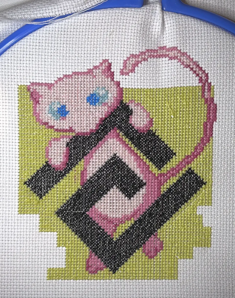 A photograph of a cross-stitch canvas. Mew, the Pokémon, is floating behind the digiLab company logo.